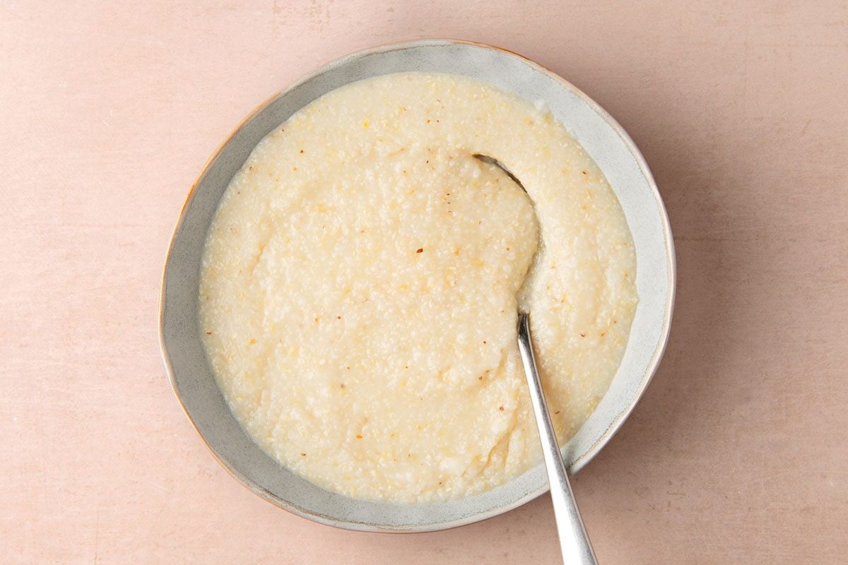 overhead shot of a bowl of creamy grits with a silver spoon lying on top of the grits, the spoon has a curve in it; the bowl is light grey with white streaks in the glaze and a dark grey rim; the bowl sits on a plain light pink surface