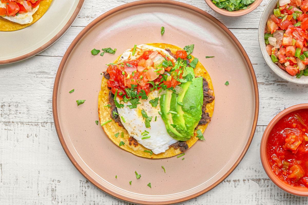 overhead shot of Huevos Rancheros is served on a brown plate, with a fried egg on top of a tortilla with refried beans and avocado slices, and topped with salsa and cilantro, The dish is surrounded by other bowls, each containing different ingredients for the Huevos Rancheros, including salsa, chopped cilantro, and a red sauce; The background is a light wooden table