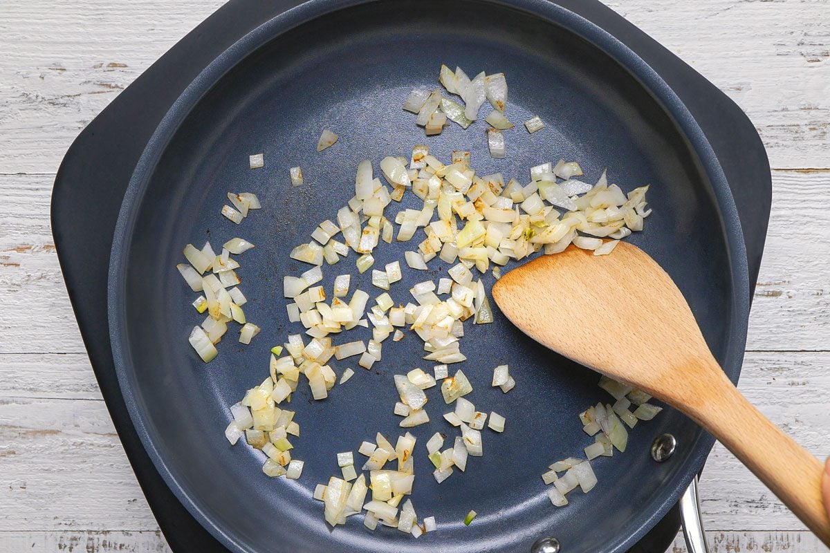 overhead shot of a black frying pan with a wooden spatula in it, the pan is placed on a white wooden surface; the pan has diced onions that have been cooked in it