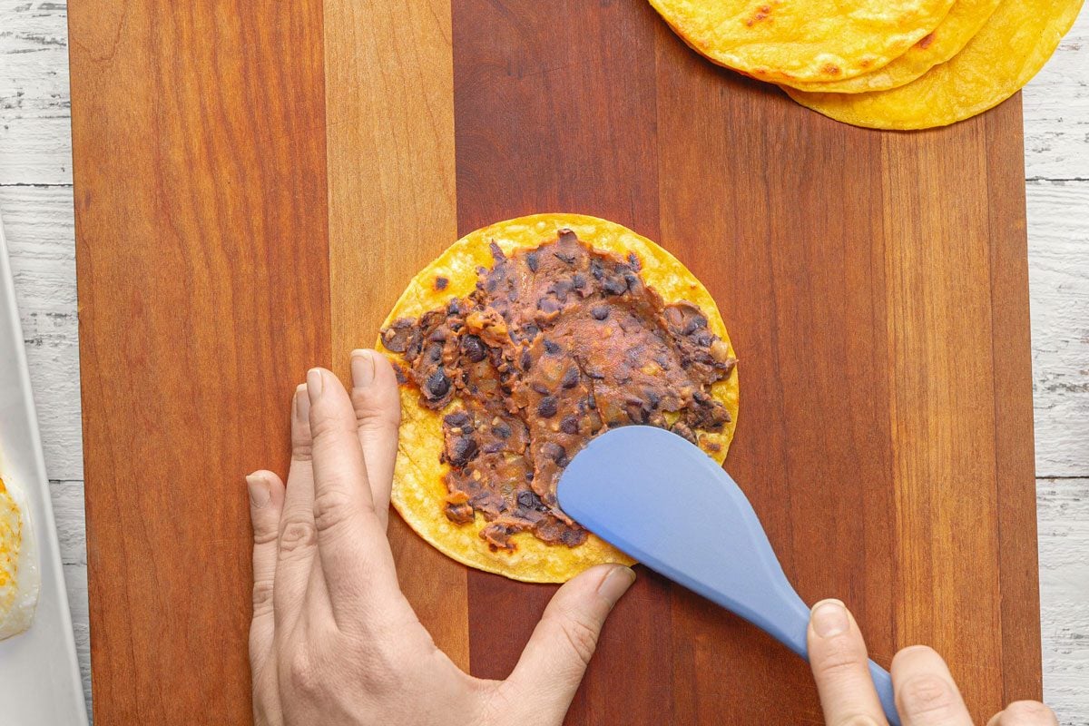 overhead shot of a person spreading black bean refried beans on a corn tortilla with a blue spatula; the tortilla is on a wooden cutting board; there are two other tortillas on the cutting board, as well as a white plate with fried eggs and a glass bowl filled with a bean and vegetable mixture