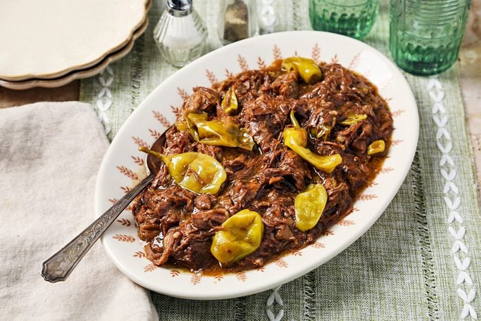 Cover push pull image of Taste of Home's Instant Pot Mississippi Pot Roast shot on a brown surface with a green linen. Pot roast shredded with peppers on a serving dish.