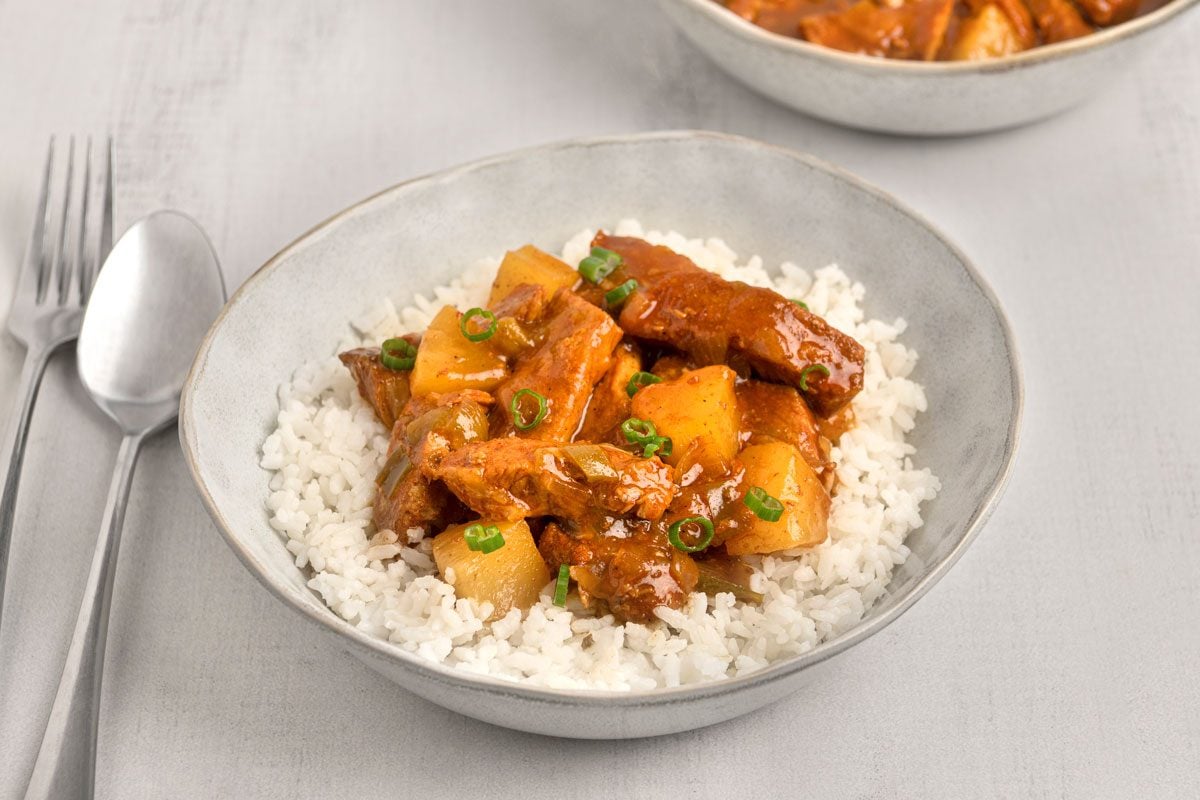 3/4th shot of a bowl of Instant pot pork loin served over white rice, a spoon and fork are placed to the left of the bowl on a light gray surface, another bowl of the same dish is partially visible in the background