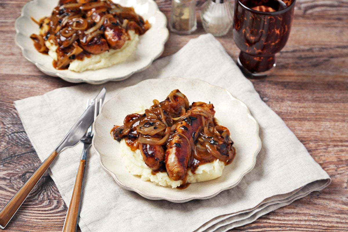 Irish Bangers and Mash shot on a brown wooden surface. Bangers and gravy served on top of mashed potatoes on scalloped plates; 3/4th