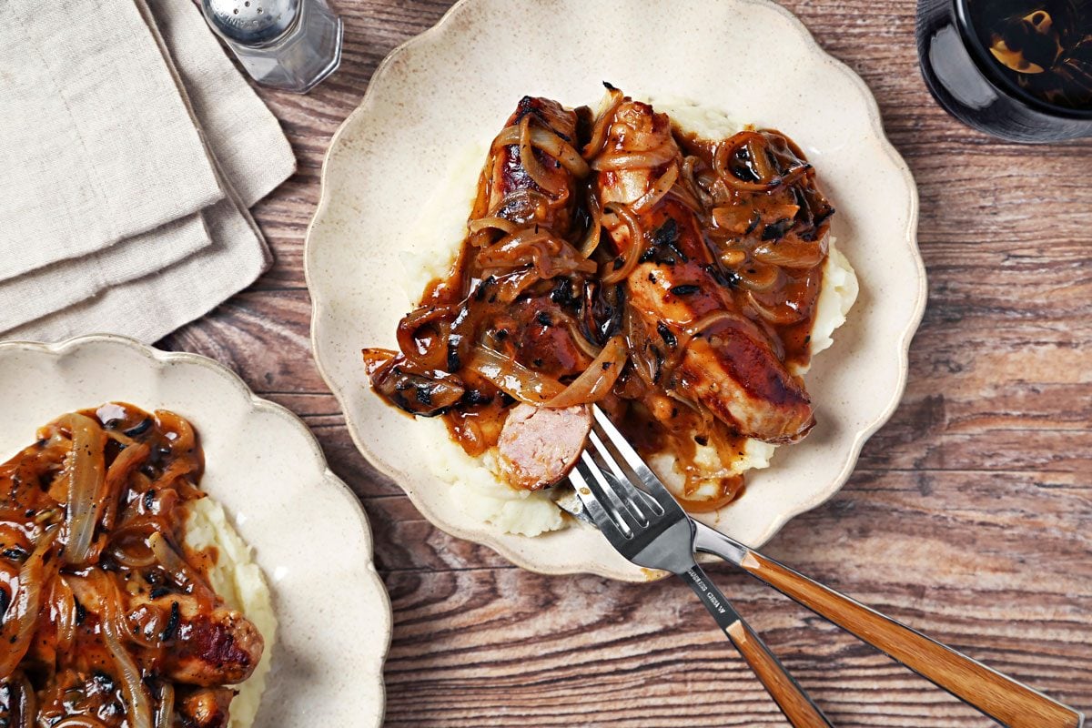 Irish Bangers and Mash shot on a brown wooden surface. Bangers and gravy served on top of mashed potatoes on scalloped plates; overhead