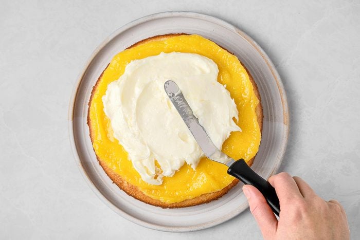 Spread cream mixture over cake kept on top of a grey marbletop.
