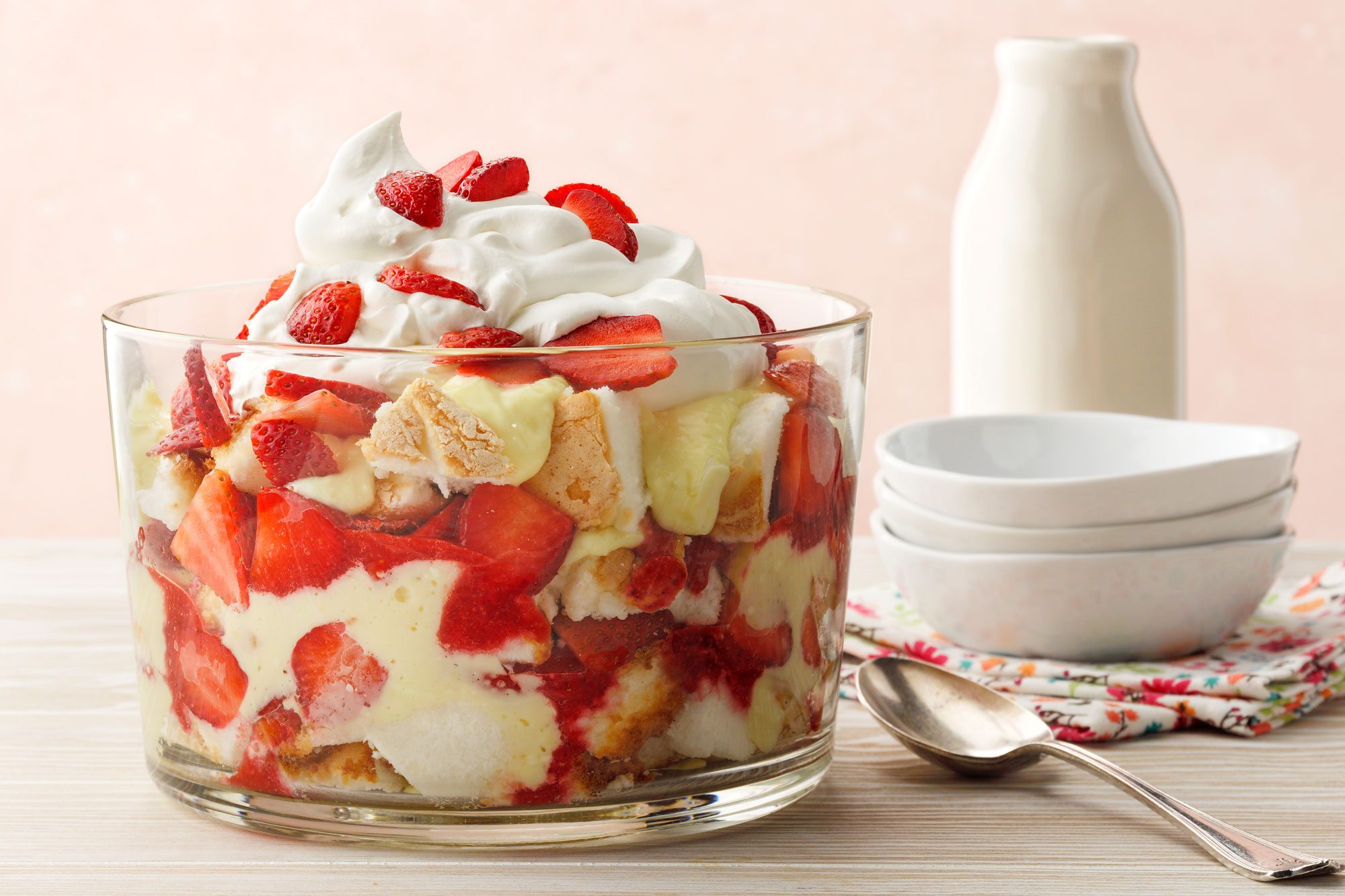 Strawberry Lemon Trifle, white washed wood surface; light wood surface; russian surface; trifle bowl; strawberries; lemon pudding; angel food cake; whipped topping; cool whip; lemon zest; milk bottle; white bowl; spoon
