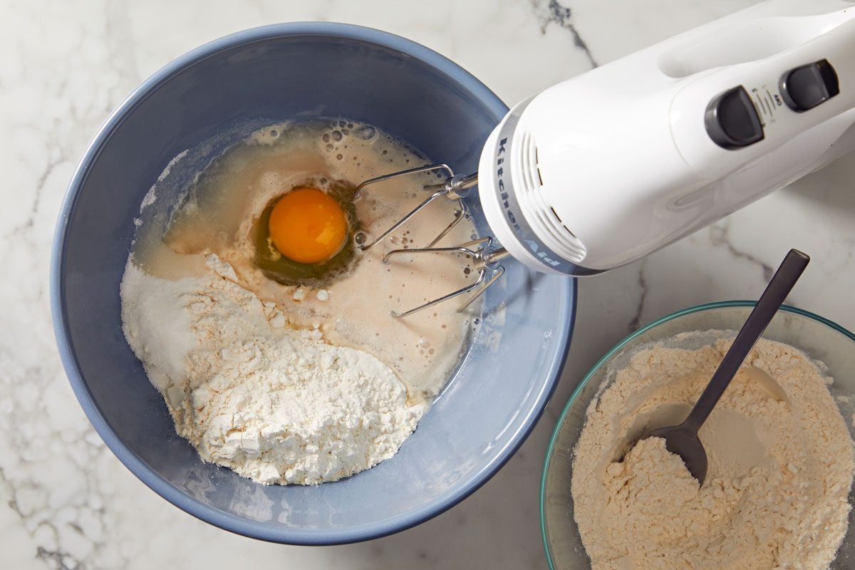 overhead shot of a mixing setup in a kitchen, a blue mixing bowl filled with flour and an egg in the mixture, a white hand mixer sits above the bowl, beside it, an additional bowl holds dry flour, with a spoon, on a marble countertop