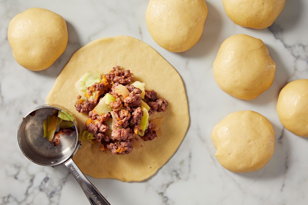 overhead shot of a round piece of dough laid flat with a mixture of ground beef, chopped vegetables, and seasoning placed in the middle; surrounding this main dough piece, several smaller balls of dough can be seen, all resting on a light marble surface; a measuring cup, positioned next to the filling