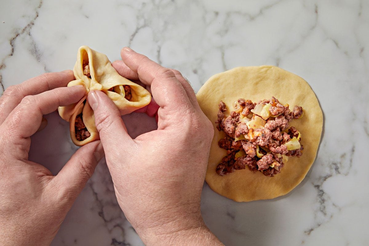 overhead shot of a pair of hands folding dough over a mixture of ground meat and vegetables on a marble countertop; the dough is partially shaped into a triangular form, next to it, there’s a larger round piece of dough topped with the same meat and vegetable filling