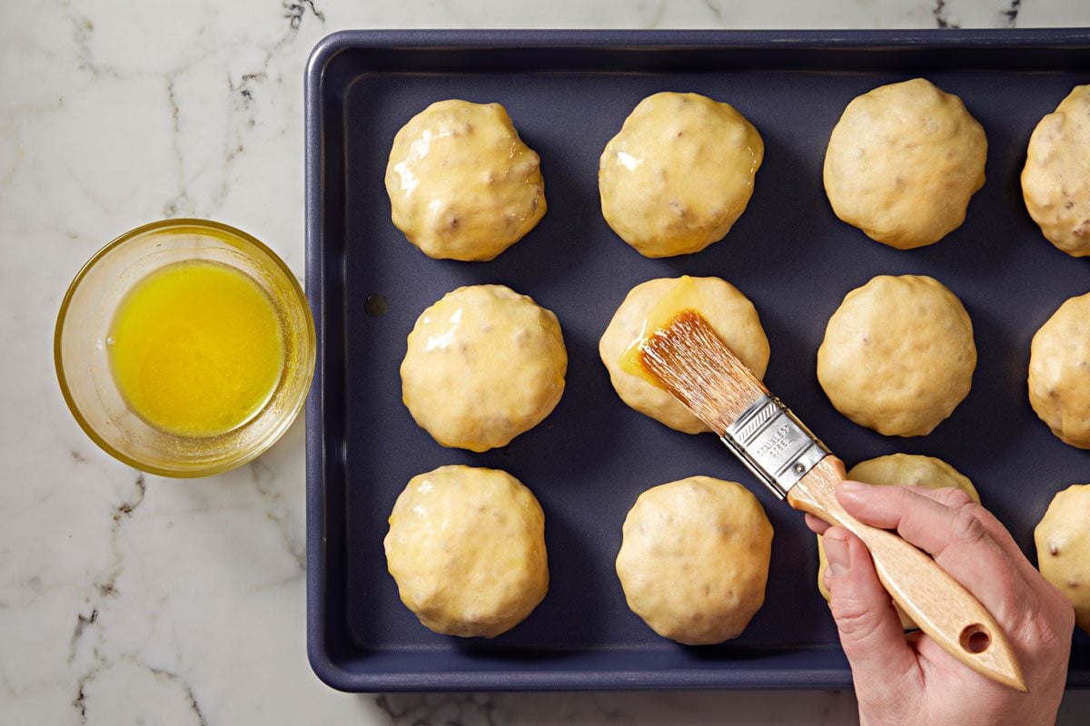 overhead shot of a baking tray filled with round dough balls; the dough is being brushed with melted butter or glaze; to the left, a small bowl containing the same liquid sits on a light marble surface