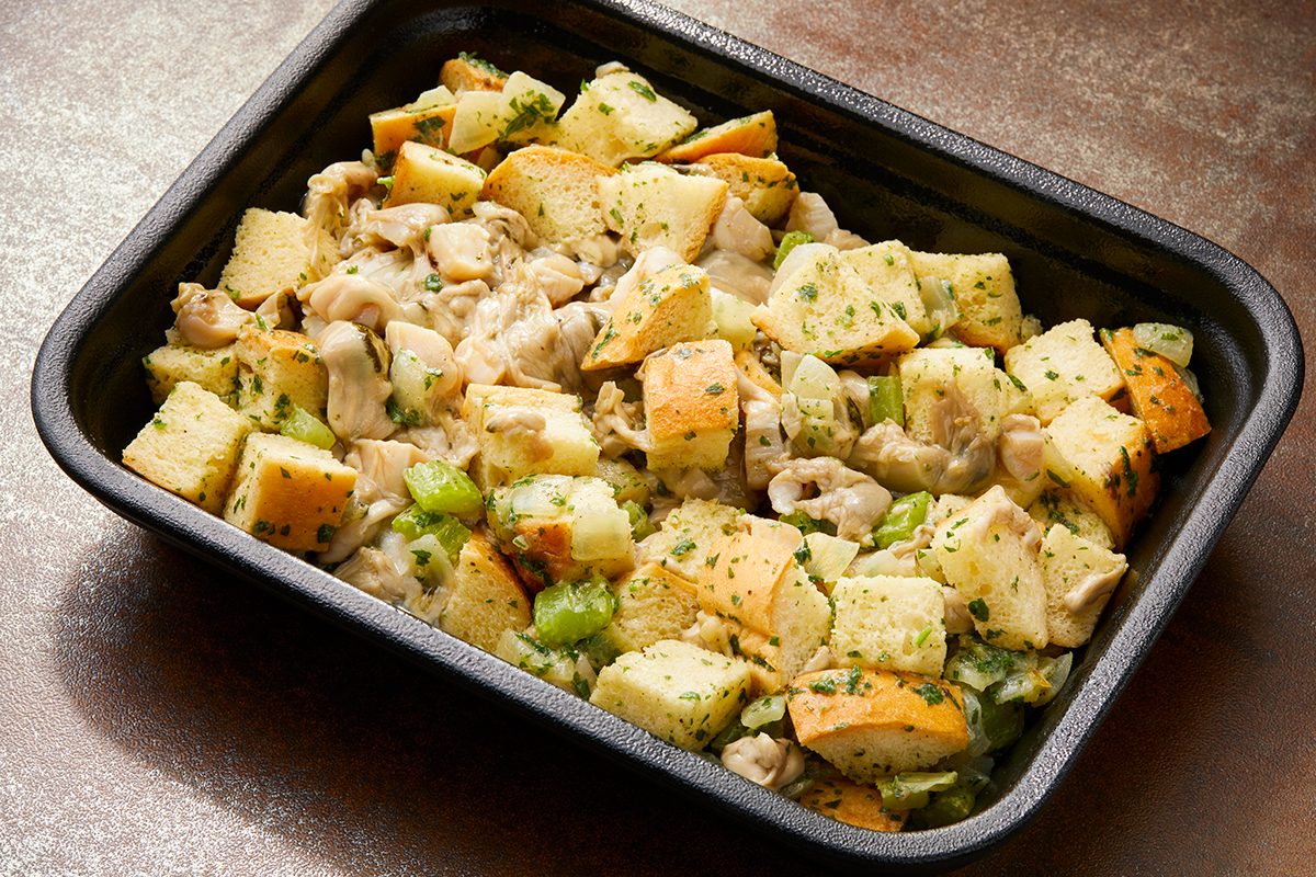 A baking tray filled with homemade stuffing. The dish contains chunks of bread, pieces of chicken, and sliced celery, all seasoned with herbs.