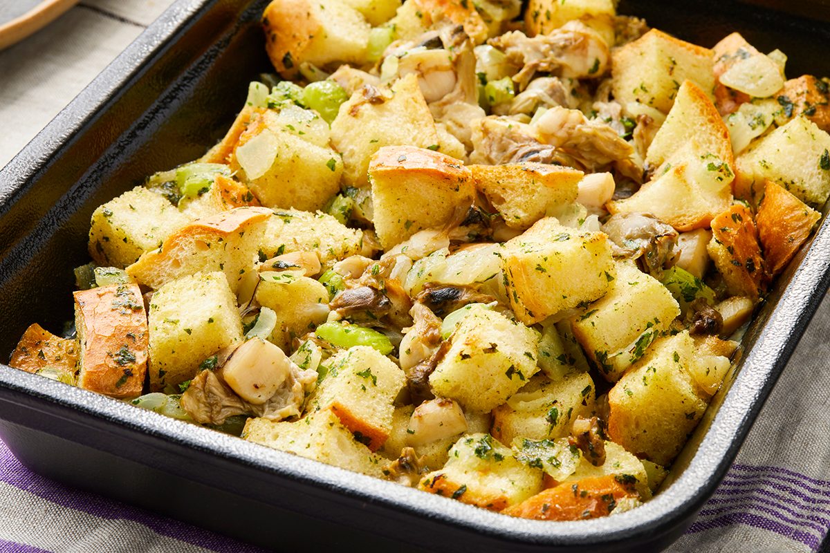 A close-up of a baking dish filled with bread stuffing. The stuffing includes cubed bread, celery, onions, and herbs. The bread appears toasted and golden, with a variety of textures visible throughout the dish.
