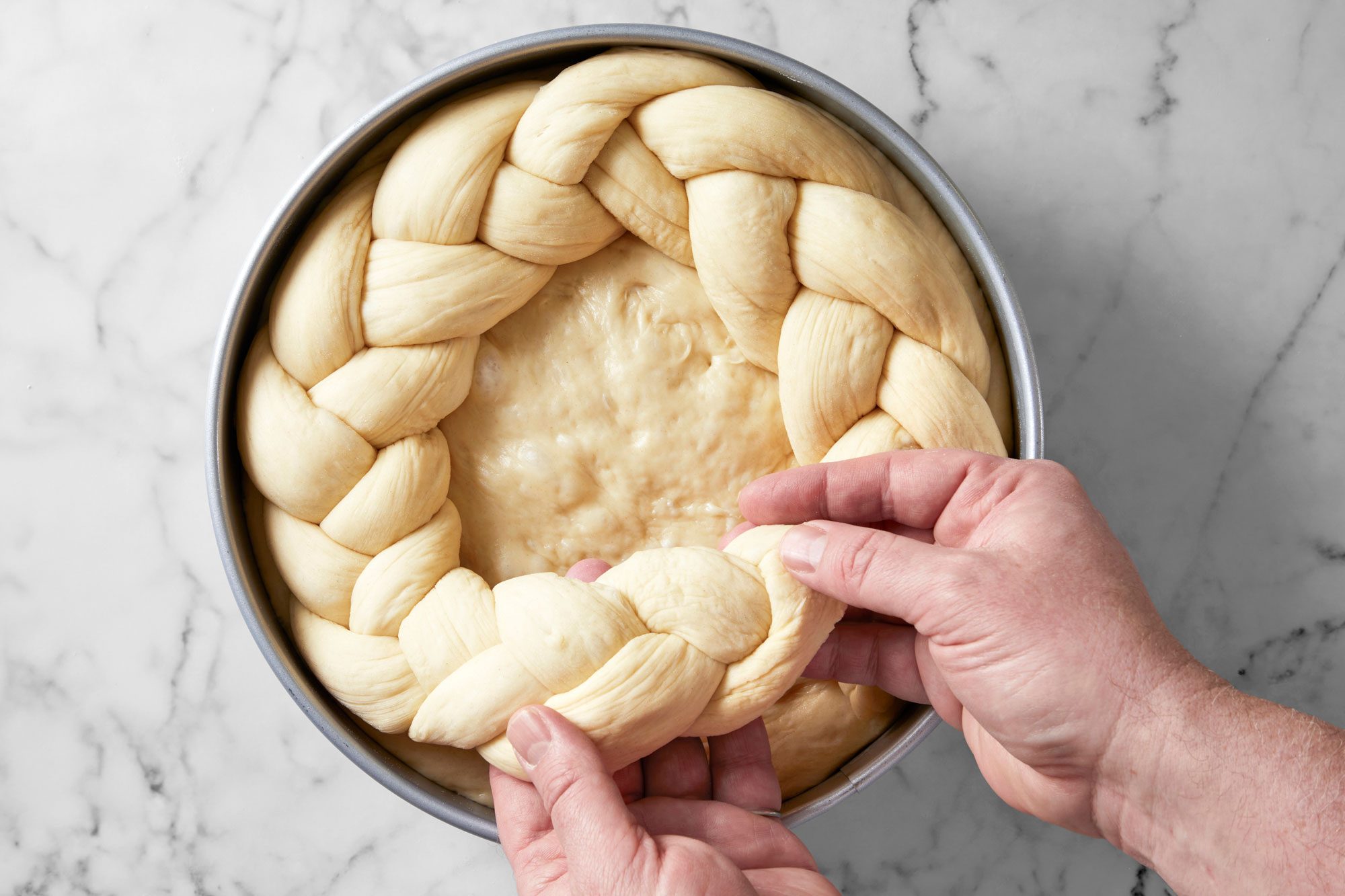 Overhead shot of place a braid around the edge of each pan; forming a circle; Trim ends of braids; reserving dough scraps; Pinch ends of braids to seal; marble surface