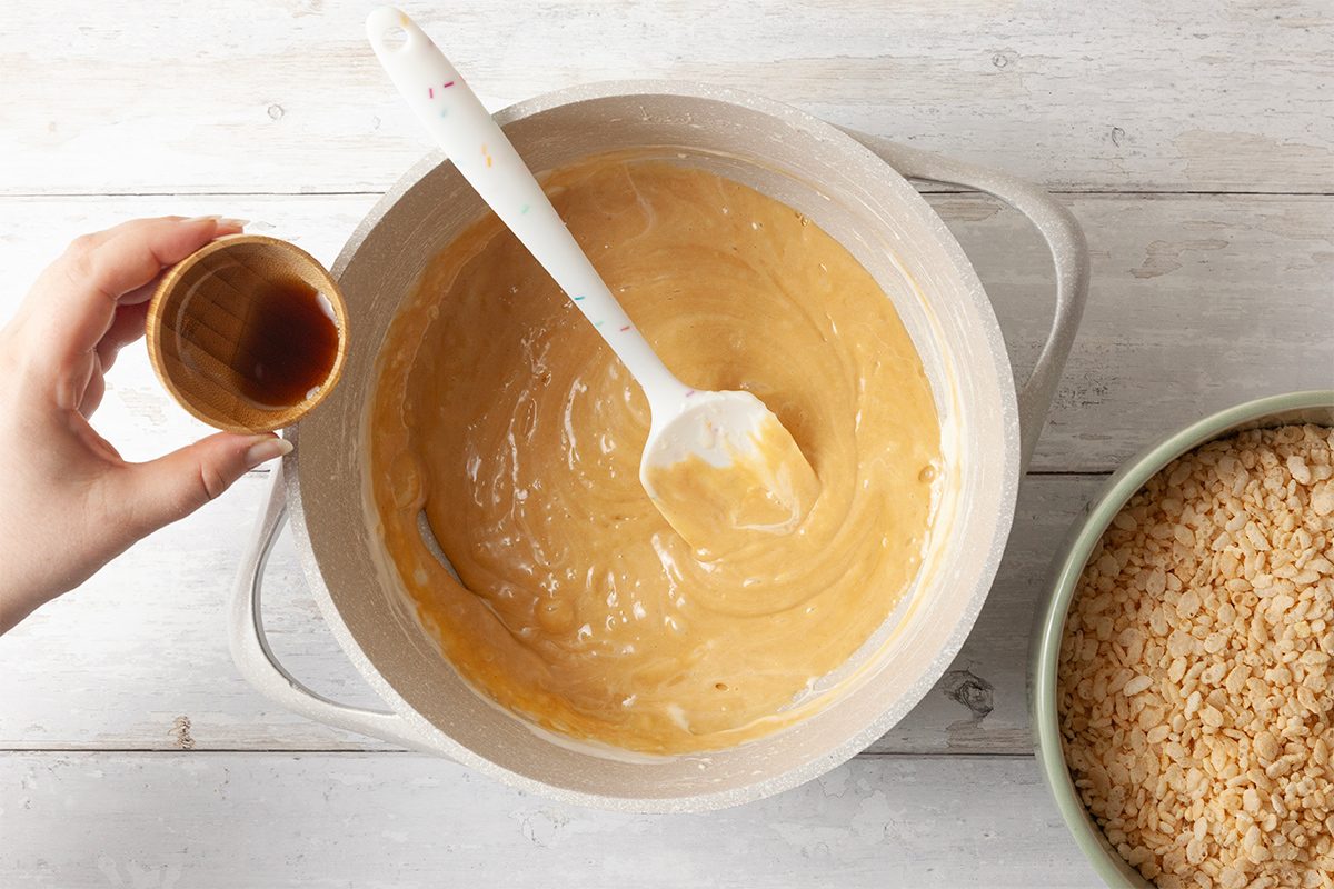 A hand holds a small cup of vanilla extract above a pot of creamy batter, which is being mixed with a white spatula. Next to the pot, there is a bowl filled with crispy rice cereal on a light wooden surface.