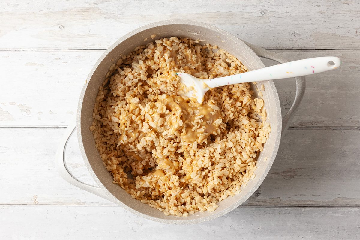 A pot filled with cooked brown rice sits on a white wooden surface. A white spoon rests in the rice, which appears fluffy and slightly compressed. The scene is simple and rustic, with neutral tones dominating the image.