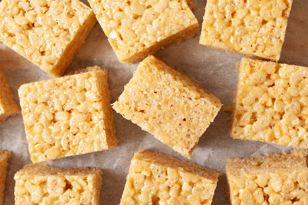 Close-up of several square rice crispy treats on wax paper. The treats have a golden, crispy texture and are aligned neatly.