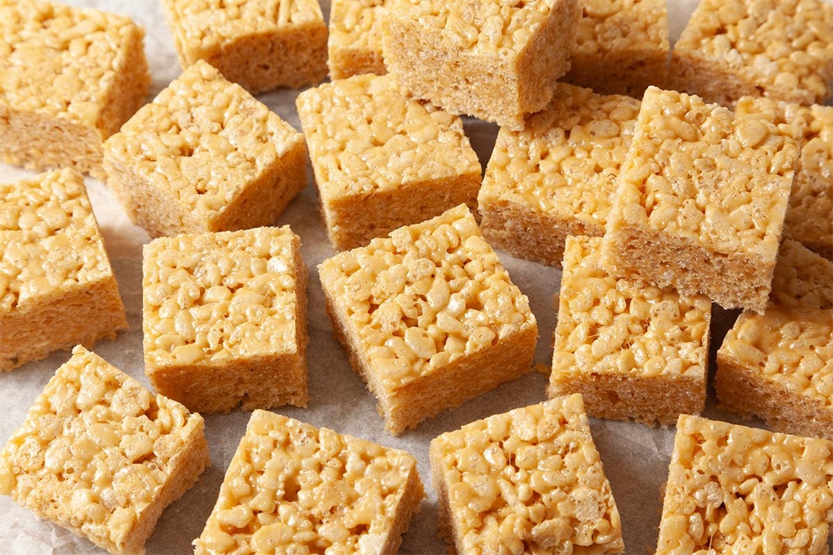 Square pieces of rice crispy treats are arranged on a white surface. They have a golden, crunchy texture from the puffed rice and marshmallow mixture, creating a sweet, chewy appearance.