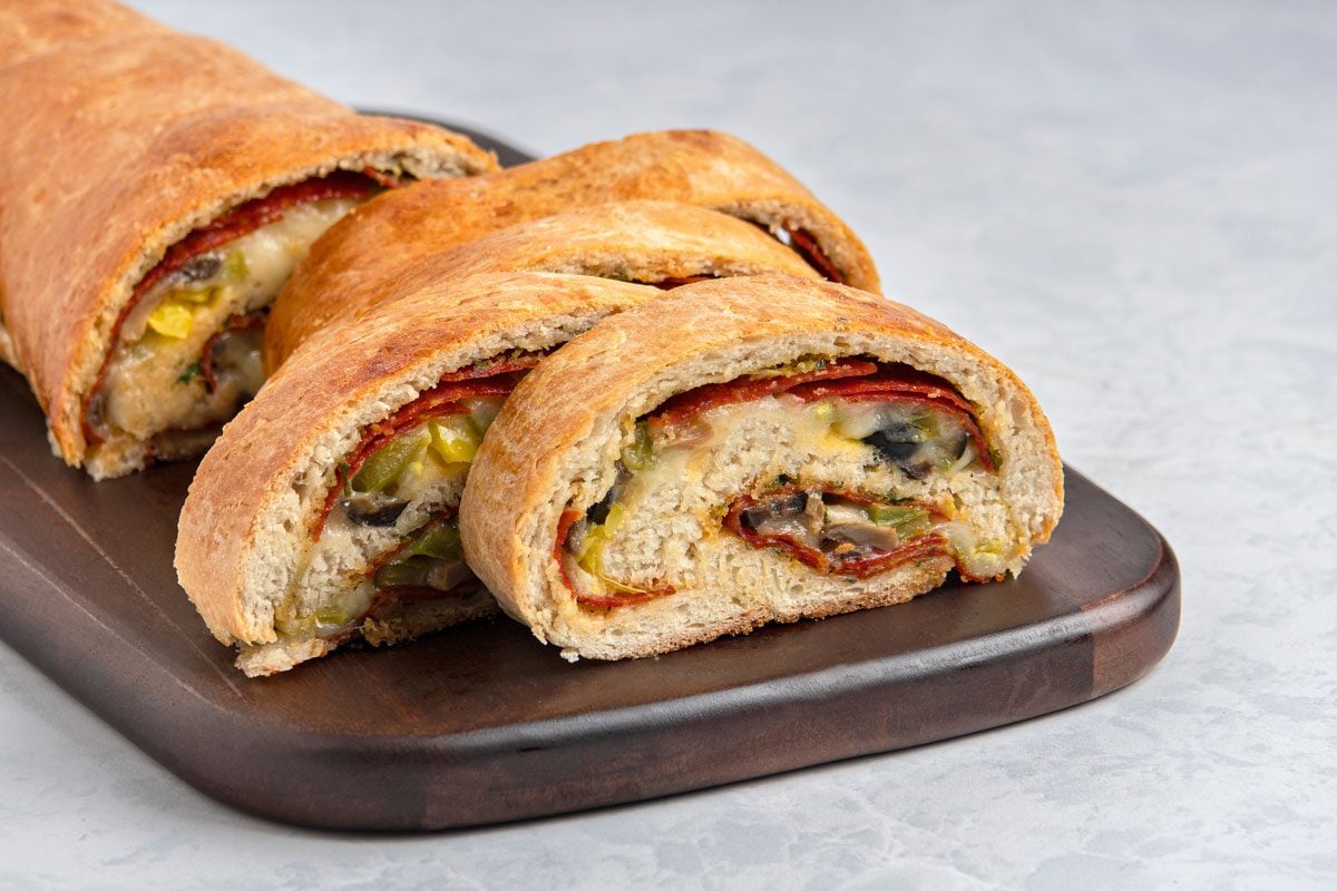 closeup shot of slices of Pepperoni Pizza Loaf arranged on a dark wooden serving board, the loaf is cut to reveal layers of dough, a light grey surface forms the background;