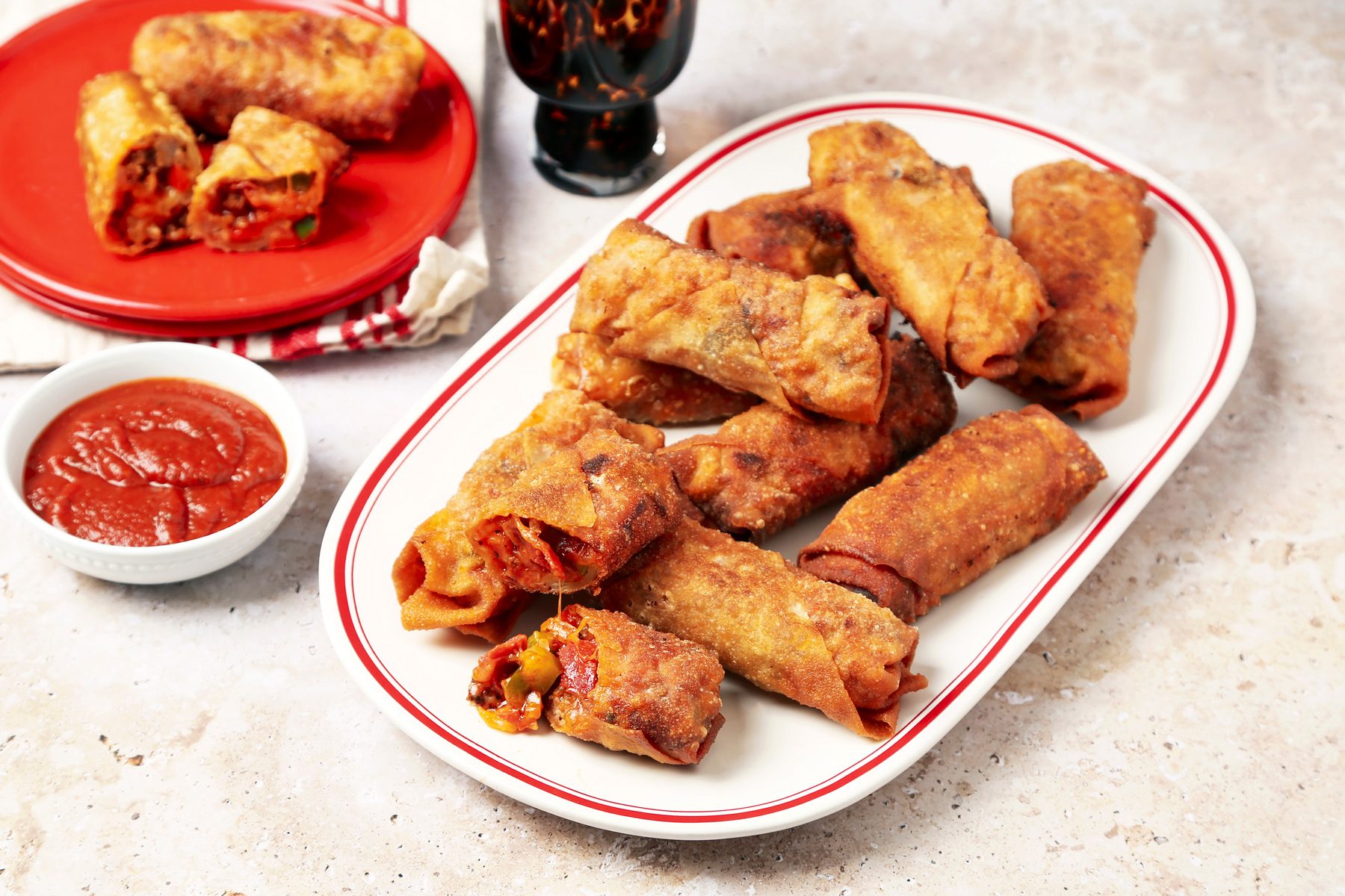 Taste of Home's Pizza Egg Rolls shot on a beige marble surface. Pizza filling inside egg roll wrappers and fried until golden brown and crispy and served with pizza sauce for dipping.