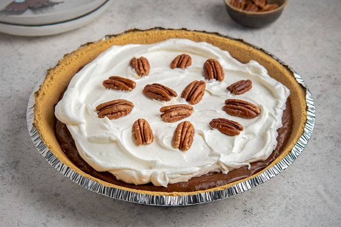 A pie in a metal dish sits on a gray surface. It has a layer of whipped cream topped with pecans, covering a chocolate filling. A small bowl with pecans is in the background.