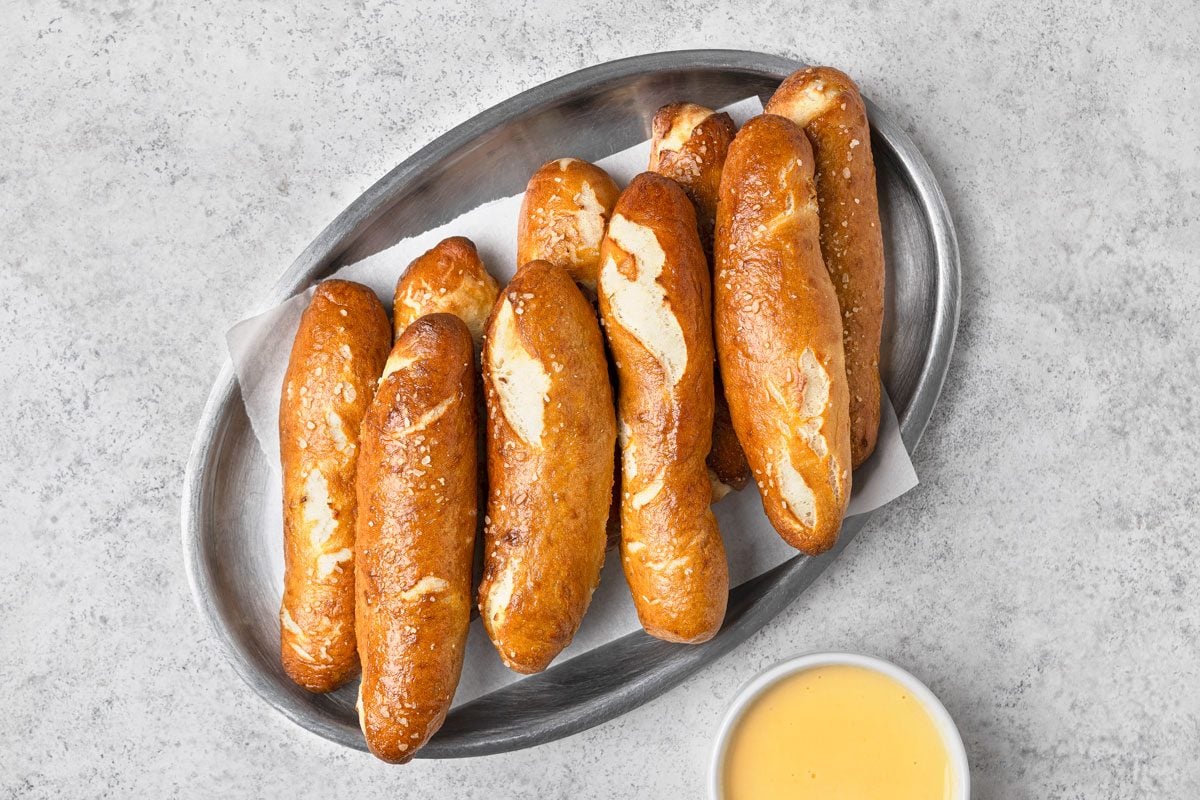 overhead shot of nine pretzel sticks arranged on a silver oval plate, they are placed on a white paper liner on the plate, and in the bottom right corner of the frame is a small white bowl filled with yellow dipping sauce, the background is a light gray surface;