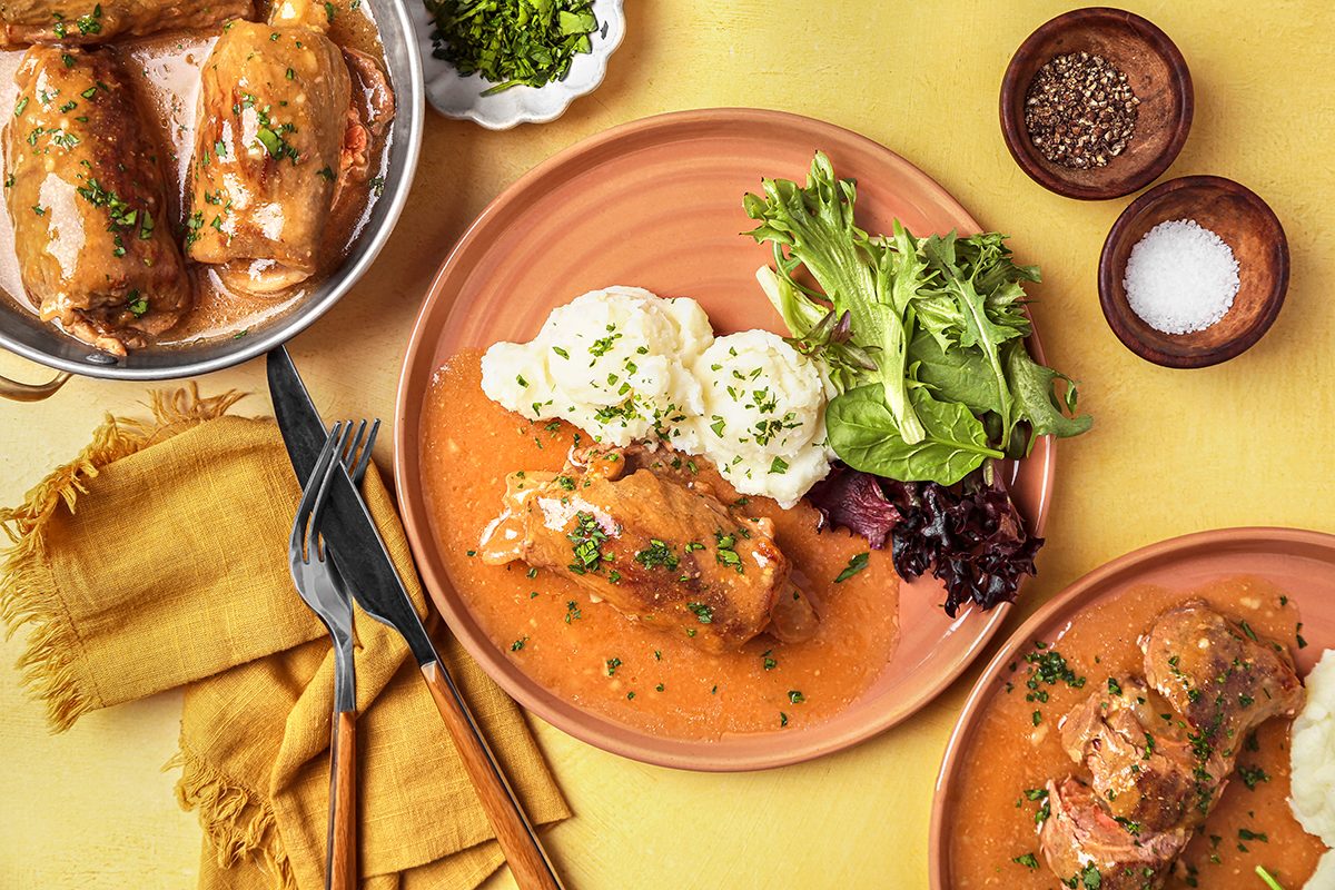 A meal featuring cabbage rolls with tomato sauce and herbs, served on a plate with mashed potatoes and a side salad. Accompanied by bowls of seasonings and garnished with fresh parsley. Yellow napkins and utensils are placed beside the dish.