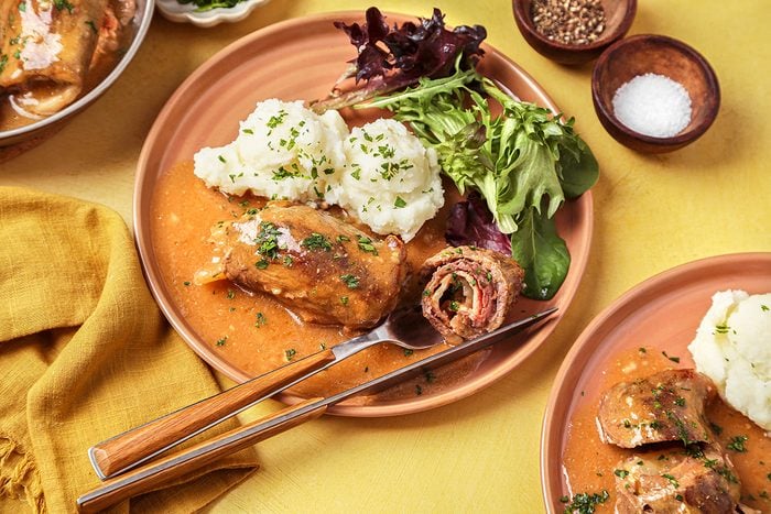 A plate of stuffed cabbage rolls with tomato sauce is served alongside mashed potatoes and mixed greens. A fork and knife are placed on the plate, and small bowls of seasoning and salt are in the background.