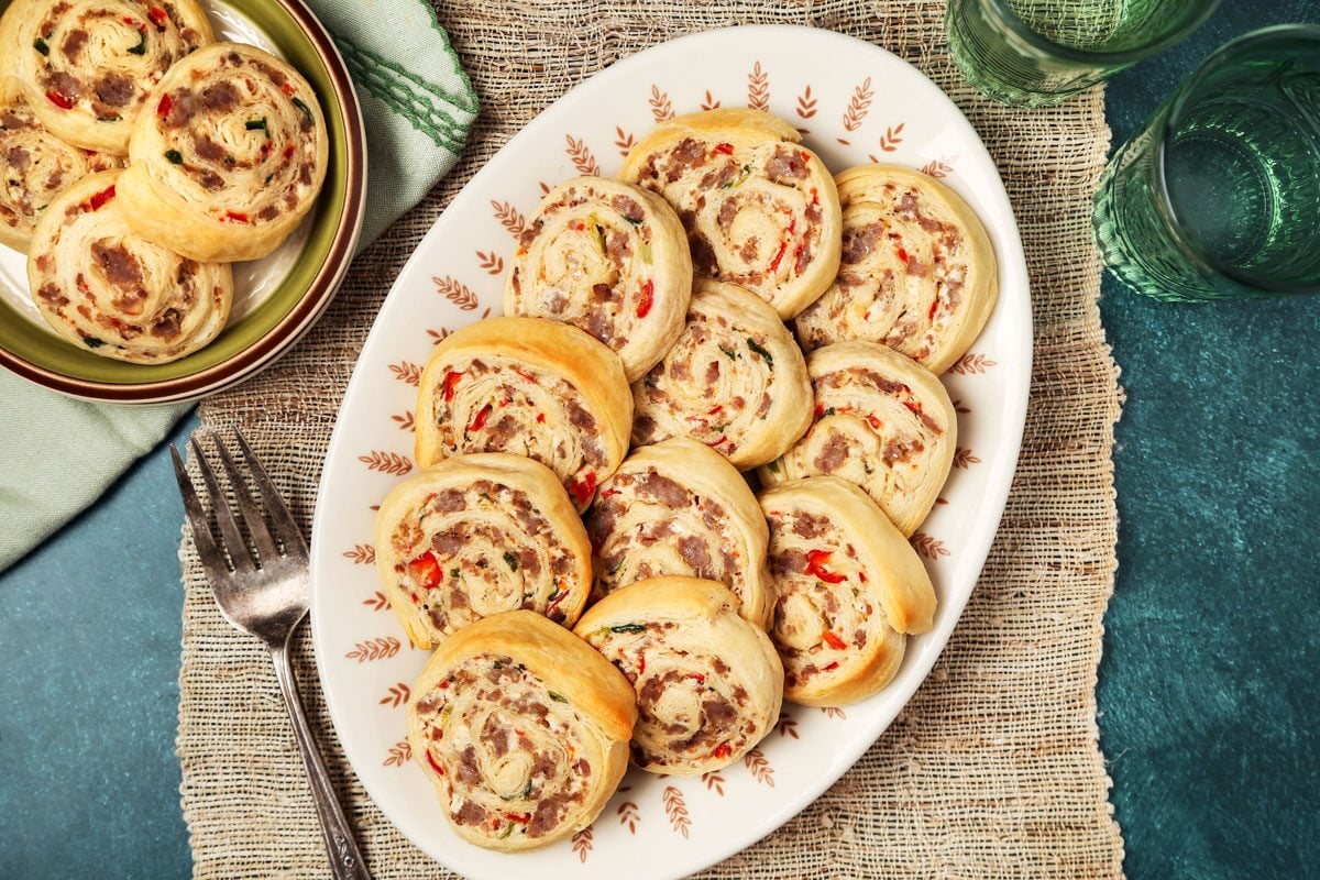 overhead shot of a plate of sausage rolls; the rolls are arranged in a circle on the plate, which is a white ceramic with a brown and white pattern; the plate is sitting on a brown placemat, which is made of woven straw; a fork is placed next to the plate; in the background, there are two green glass tumblers, one full and the other empty, sitting on a blue surface