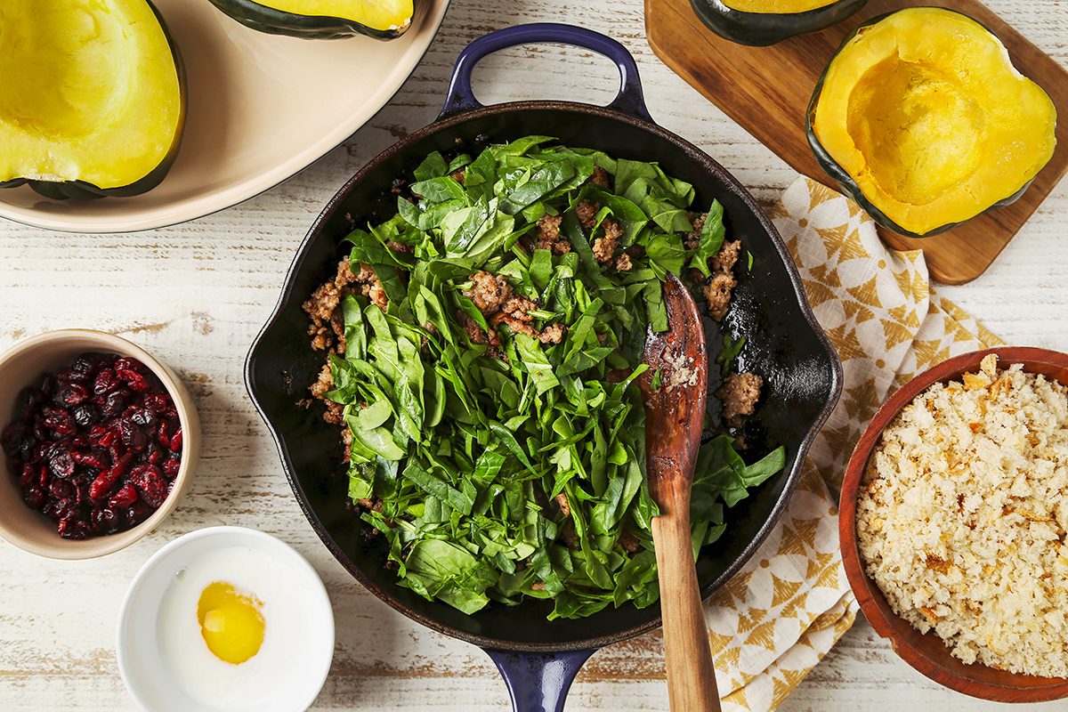 A skillet with spinach and ground meat mixture being stirred by a wooden spoon. Surrounding the skillet are acorn squash halves, a bowl of dried cranberries, a bowl of cooked rice, and a small dish with a cracked egg.