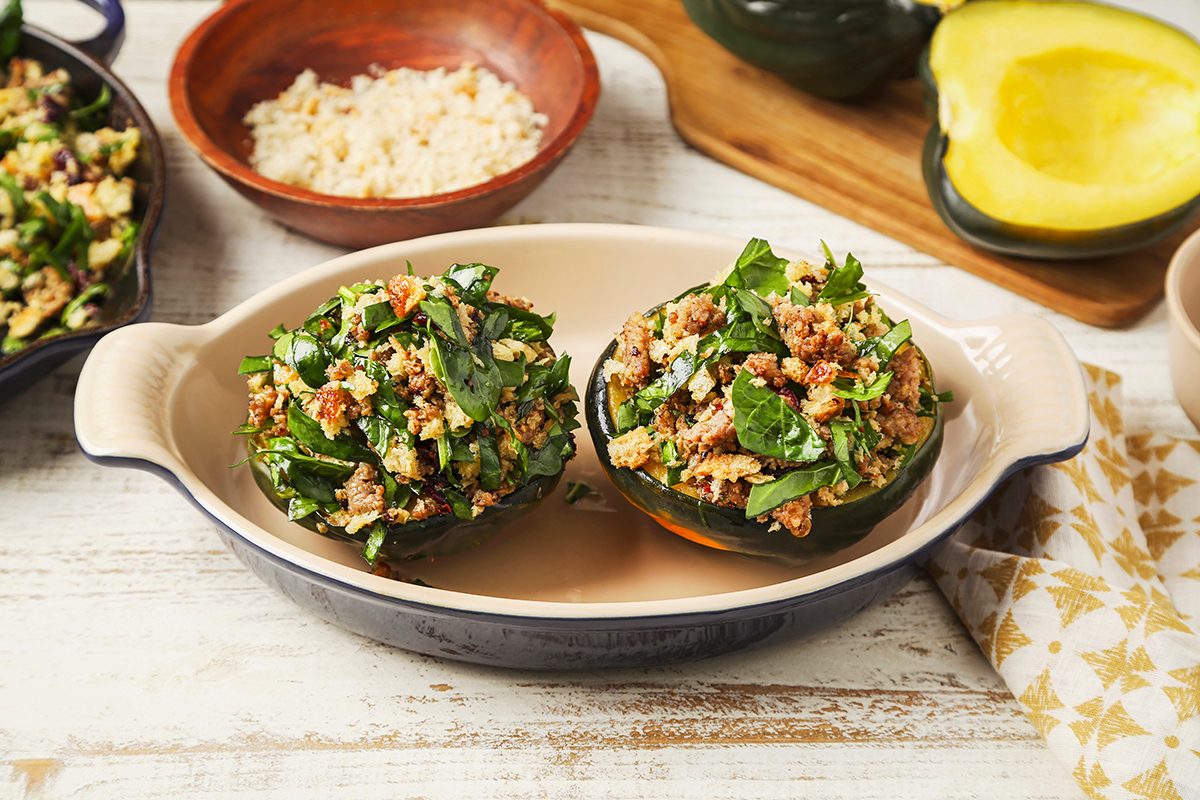Two acorn squash halves stuffed with a mixture of greens and crumbled sausage are placed in a baking dish. 