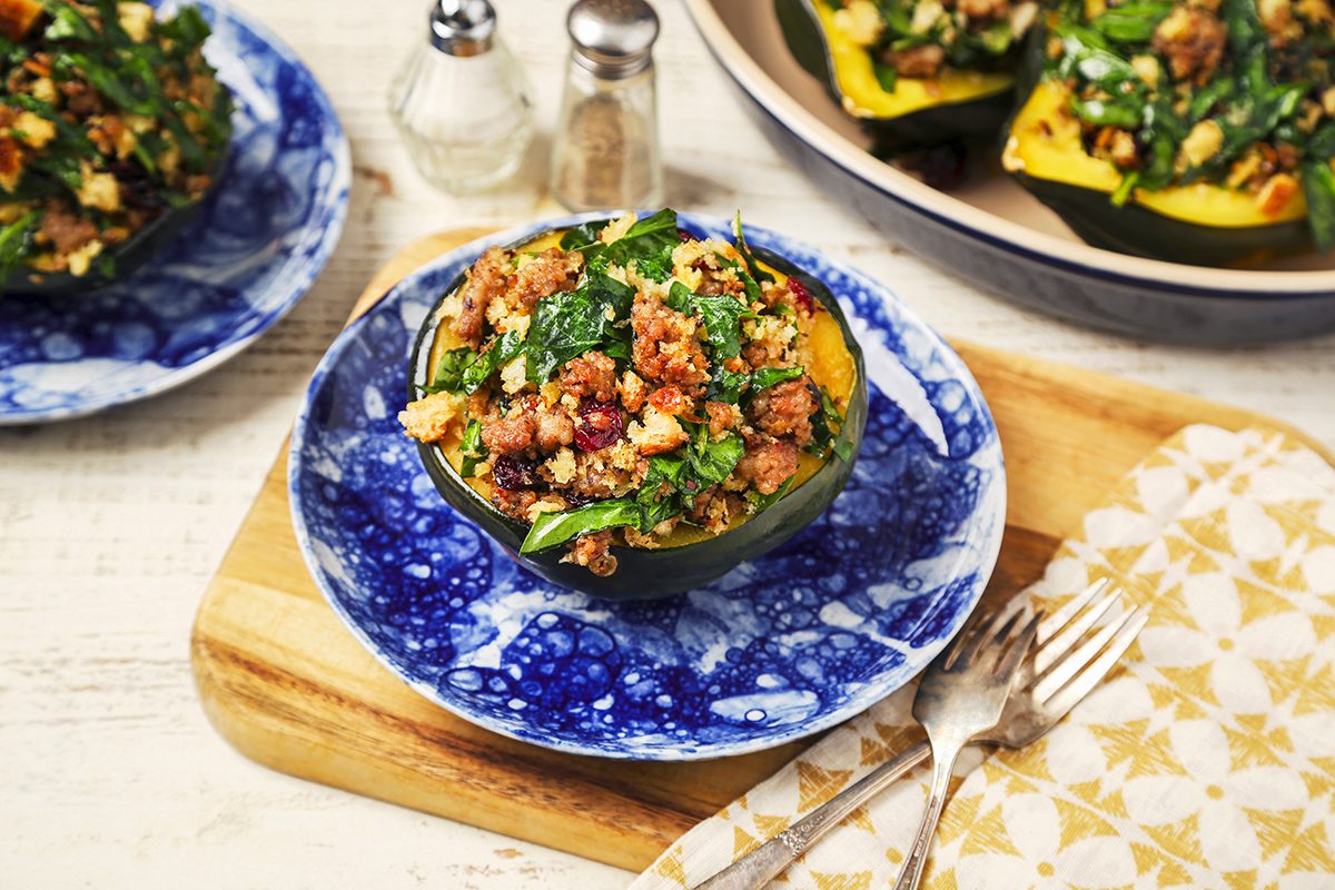 Stuffed acorn squash on a blue and white plate, filled with a mix of spinach, crumbled sausage, and grains. A wooden board beneath, a fork on a patterned napkin, and a salt and pepper shaker are in the background.