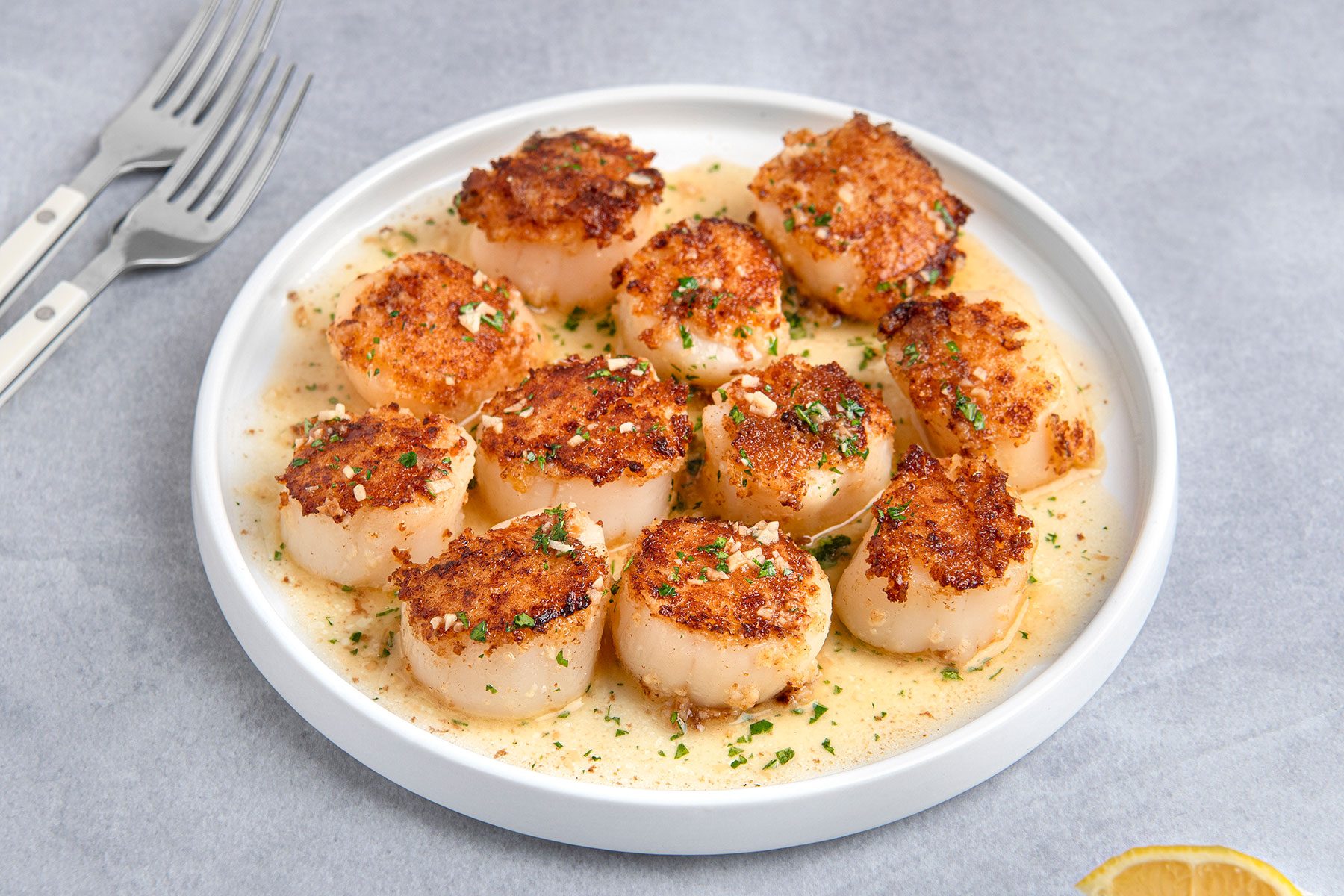 A white plate with seared scallops garnished with chopped herbs and a light sauce. Two forks rest nearby on a gray surface, and a slice of lemon is partially visible in the corner.
