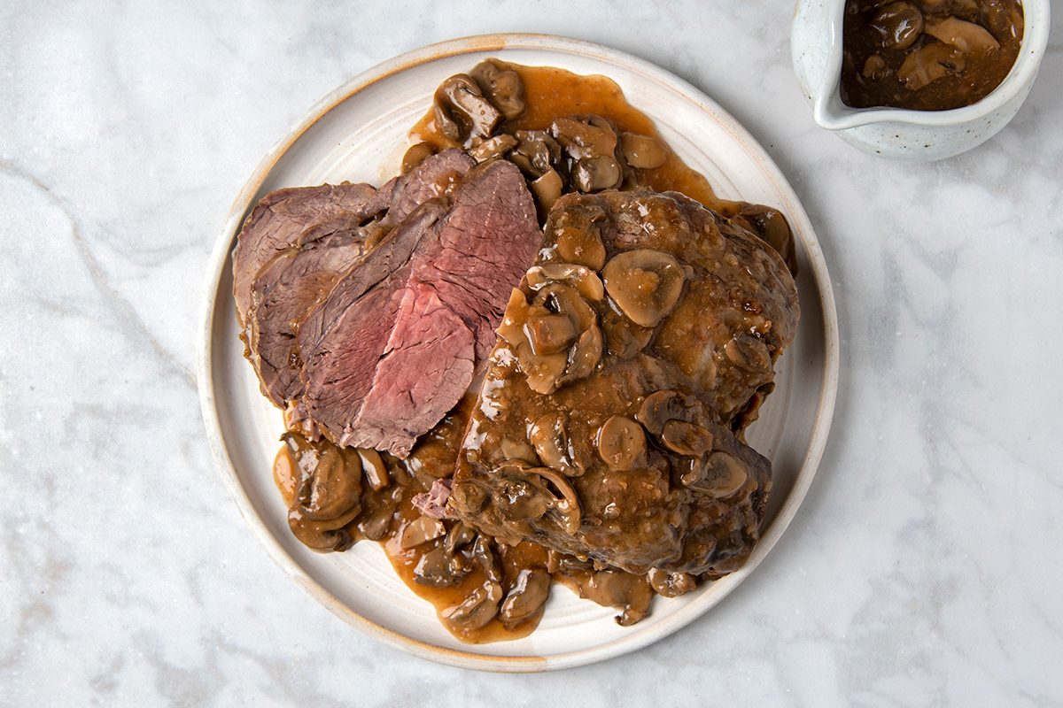 A plate of roast beef topped with sliced mushrooms and brown gravy, with a side of sauce in a white gravy boat. The beef is sliced and appears tender and juicy. The setting is on a light marble background.