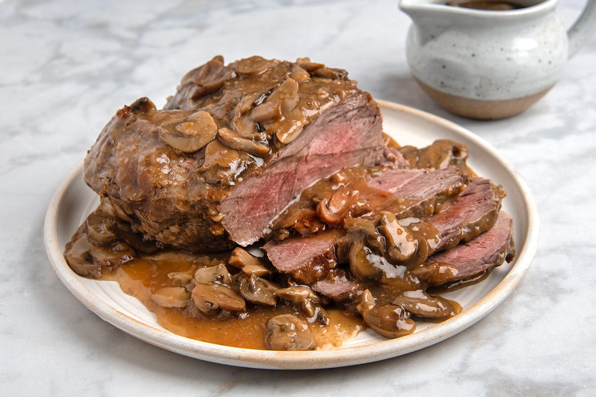 Roast beef topped with mushroom gravy, served on a white plate. The beef is sliced, revealing its juicy, tender interior. A gravy boat filled with more sauce is visible in the background. The setting is on a marble surface.