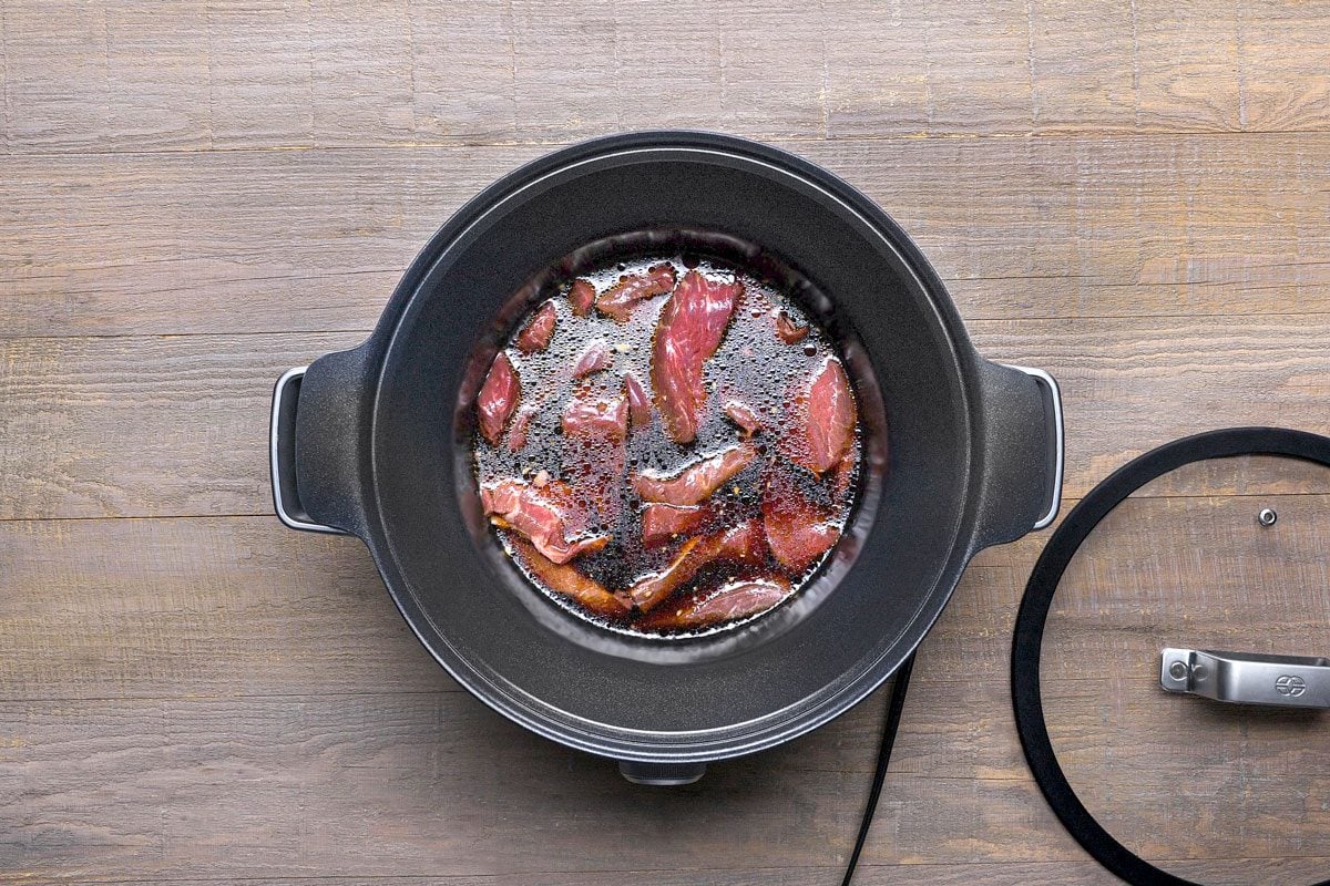 overhead shot of a black slow cooker with a glass lid on a wooden surface; the slow cooker is filled with raw meat submerged in a dark liquid
