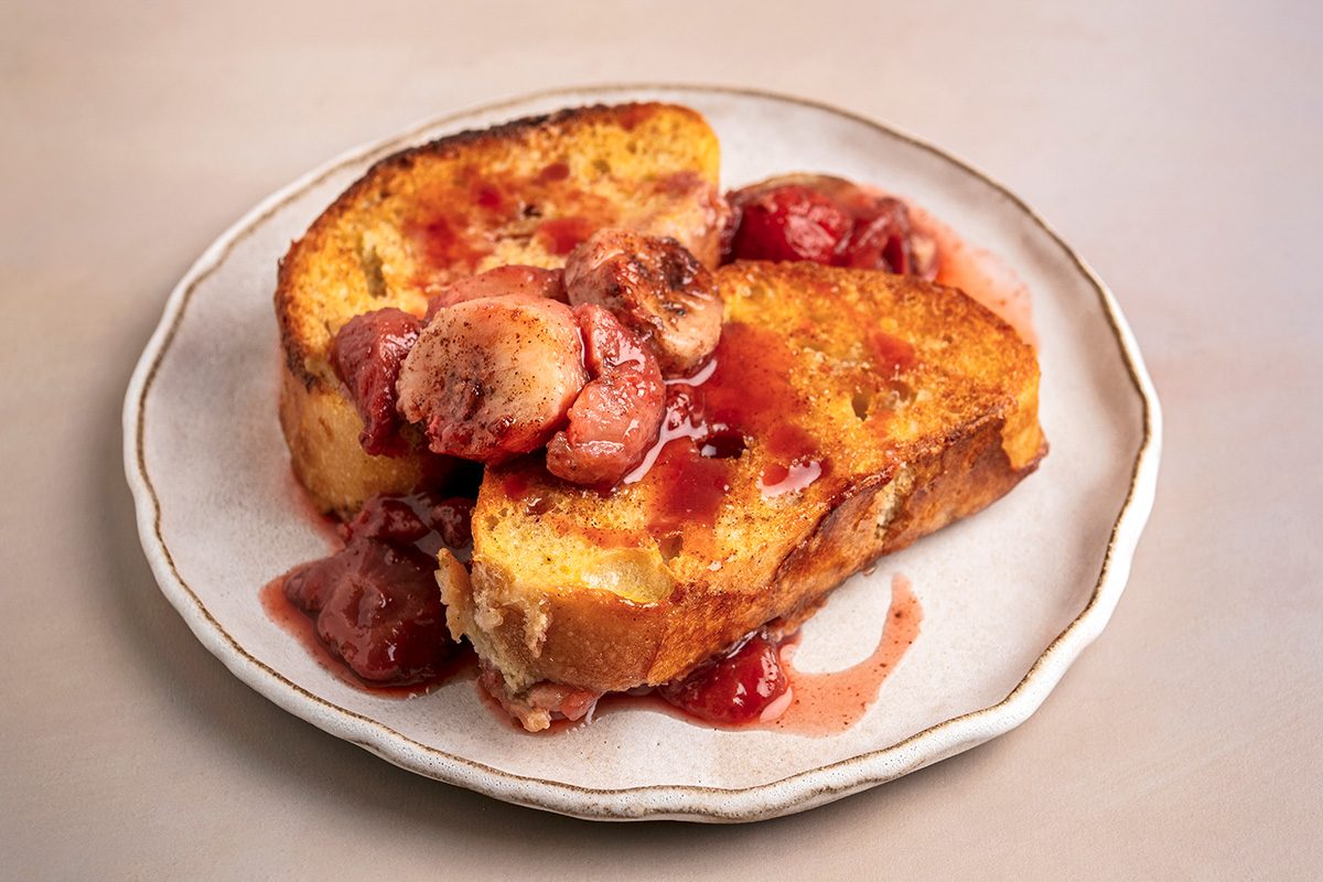 A plate with two slices of golden-brown French toast topped with glazed strawberries and syrup on a white dish with a light background.