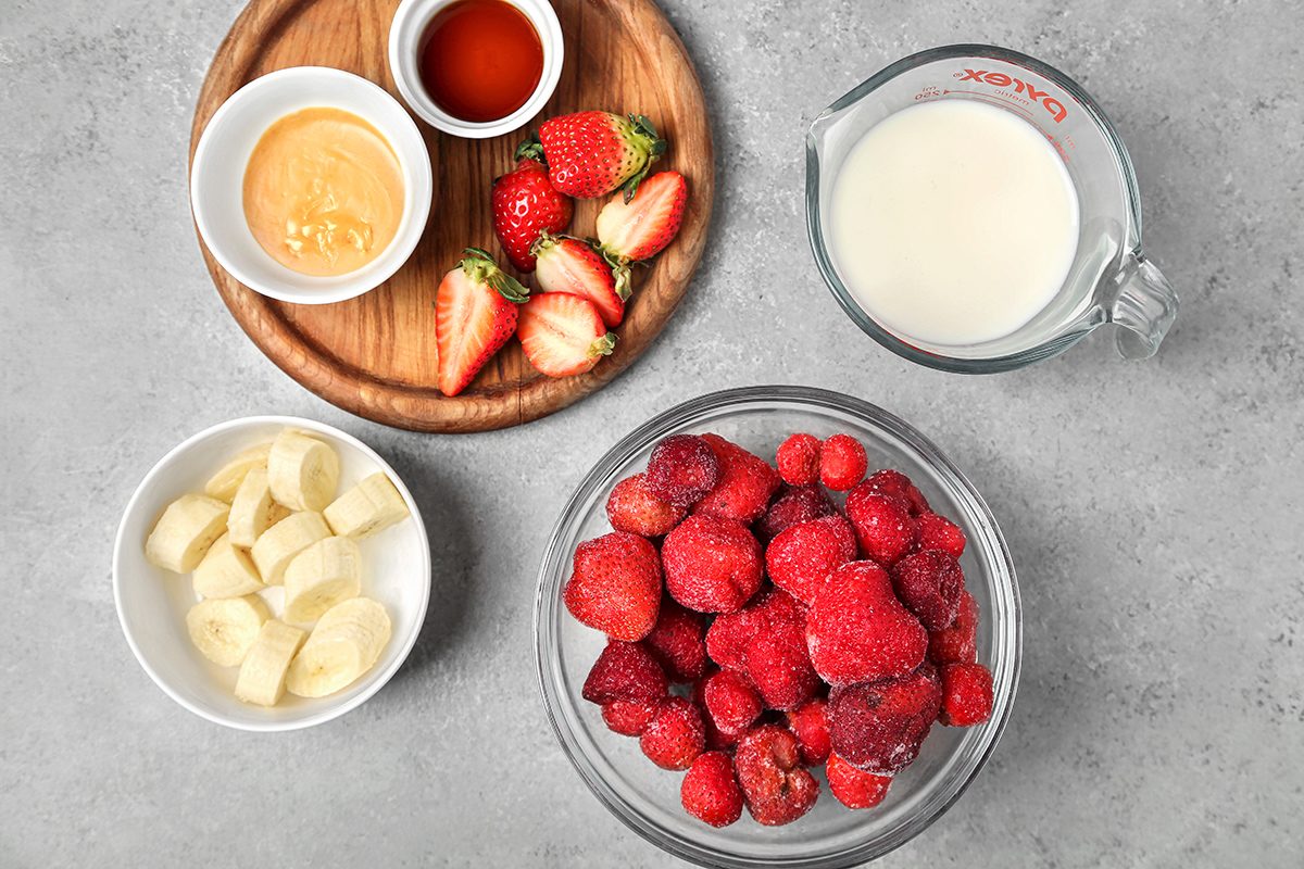 A wooden plate with honey, vanilla, and halved strawberries; a bowl with sliced bananas; a glass container with milk; and a bowl of frozen mixed berries are arranged on a gray countertop.
