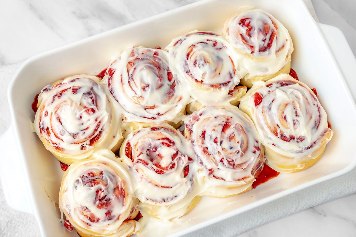 A white rectangular dish filled with frosted cinnamon rolls, swirled with a red fruity filling. The rolls are topped with a generous layer of white icing, creating a glossy appearance. The dish sits on a marble surface.