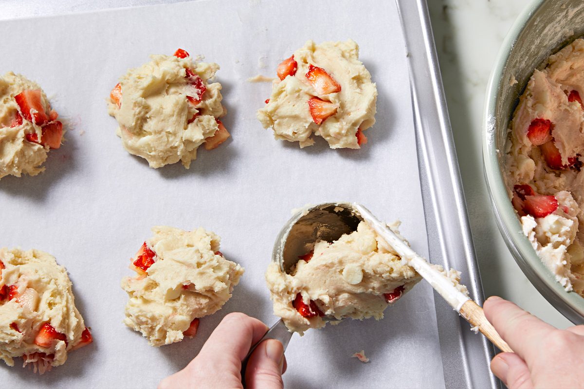 Overhead view of scooping the cookie dough onto a parchment paper-lined baking sheet using the help of a spatula. Next to it lies the large bowl containing the cookie dough.