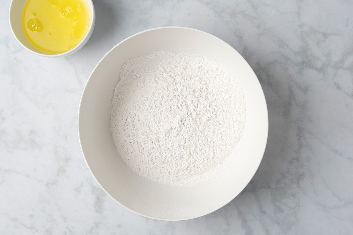A white bowl filled with flour sits on a marble countertop. To the upper left, a small white cup contains liquid egg yolk.