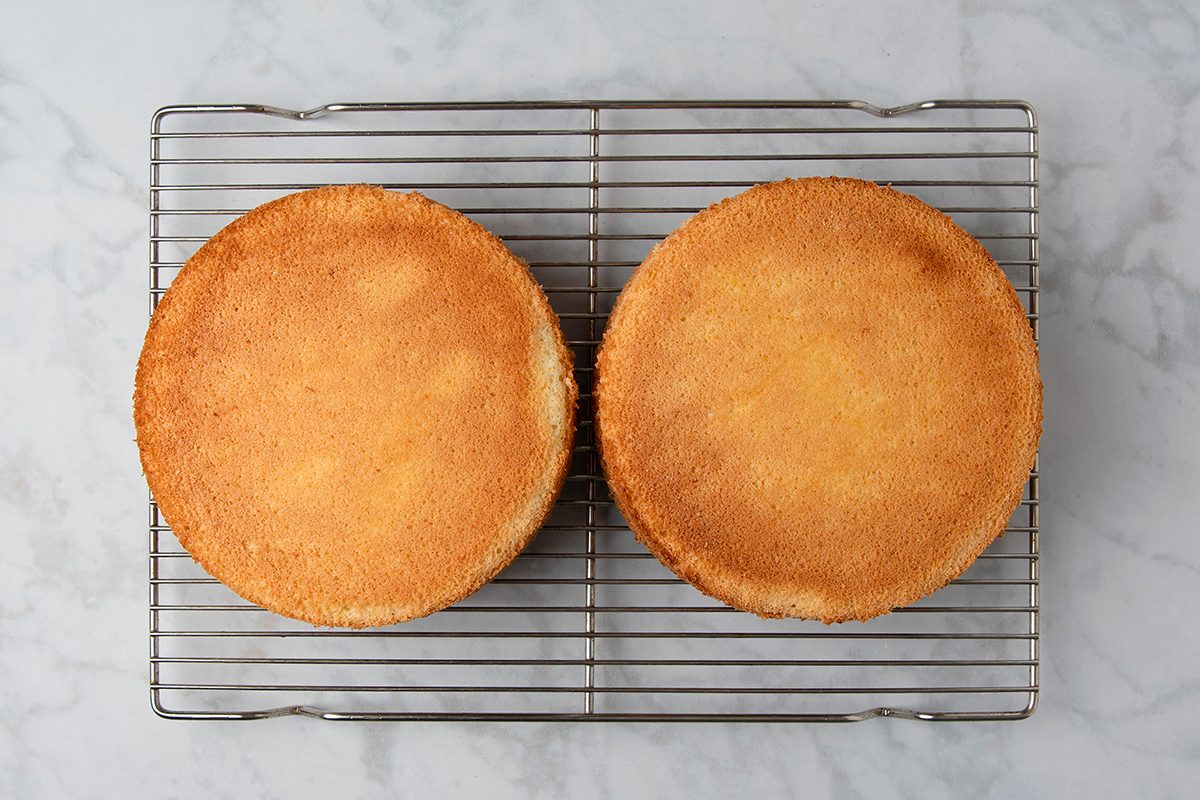 Two round sponge cakes on a wire cooling rack set on a marble surface. The cakes are golden brown and plain, without any frosting or decoration.