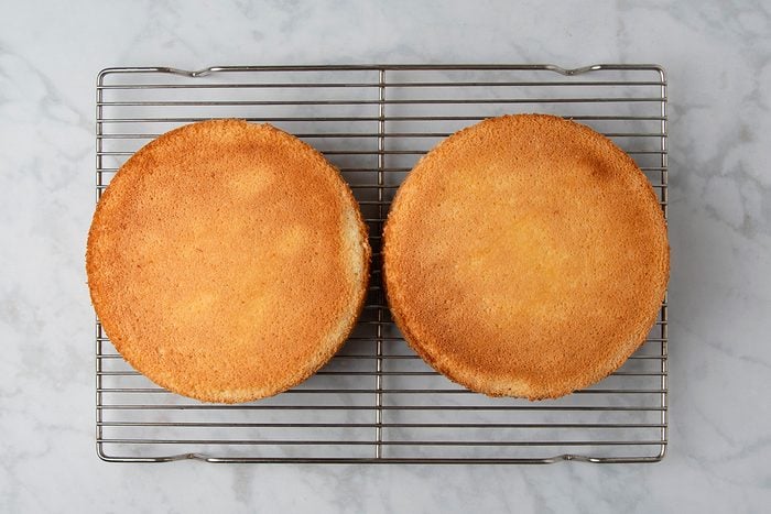 Two round sponge cakes on a wire cooling rack set on a marble surface. The cakes are golden brown and plain, without any frosting or decoration.