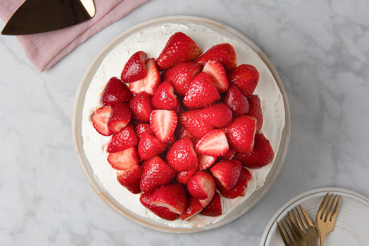 A round cake topped with a generous layer of fresh, whole strawberries on a white plate. The cake sits on a marble surface with a cake server and pink napkin nearby, and forks on a small plate to the side.