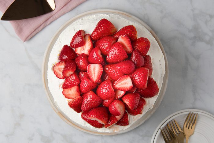 A round cake topped with a generous layer of fresh, whole strawberries on a white plate. The cake sits on a marble surface with a cake server and pink napkin nearby, and forks on a small plate to the side.