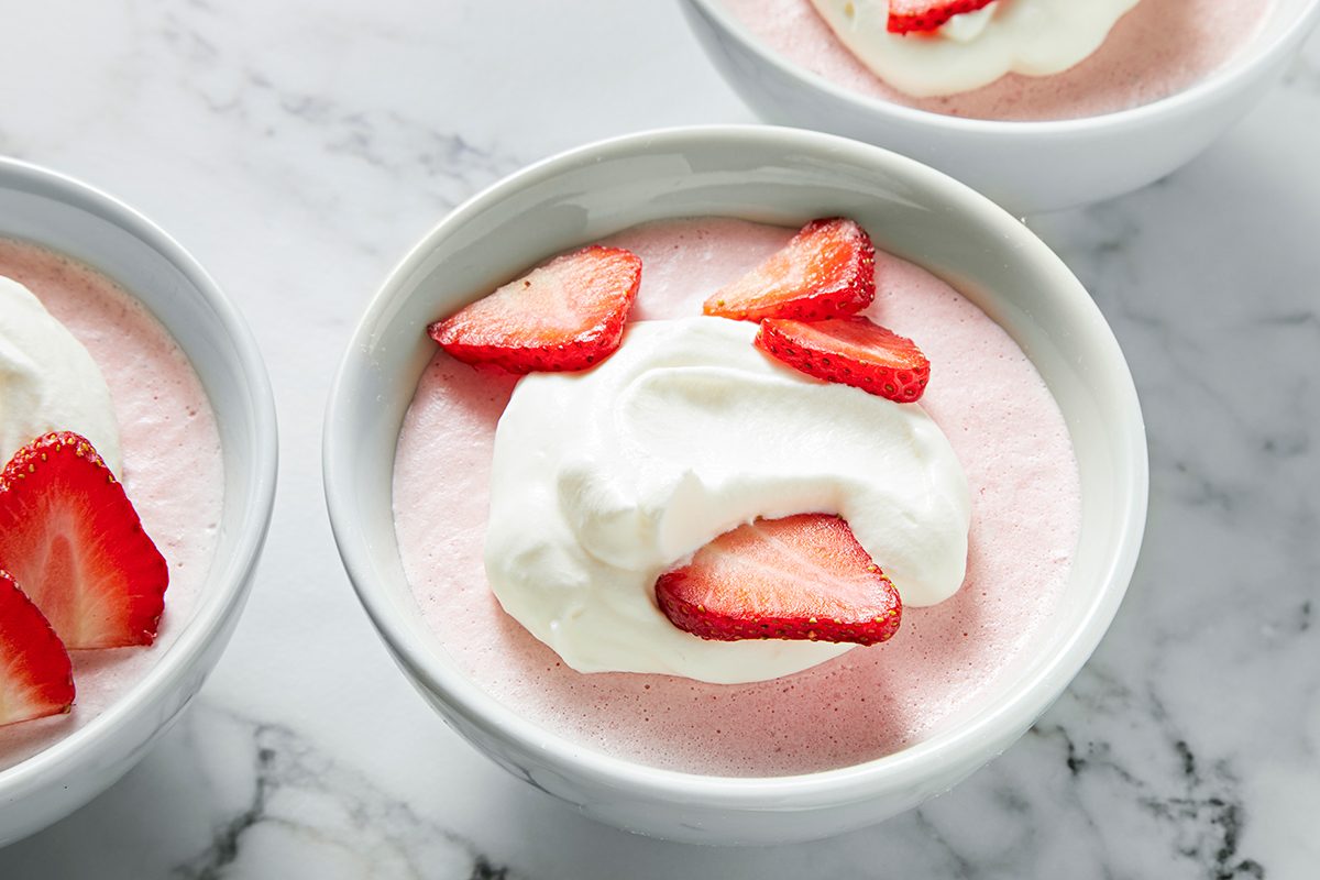 A bowl of pink mousse topped with whipped cream and sliced strawberries sits on a marble surface. Two similar bowls are partially visible on the sides.