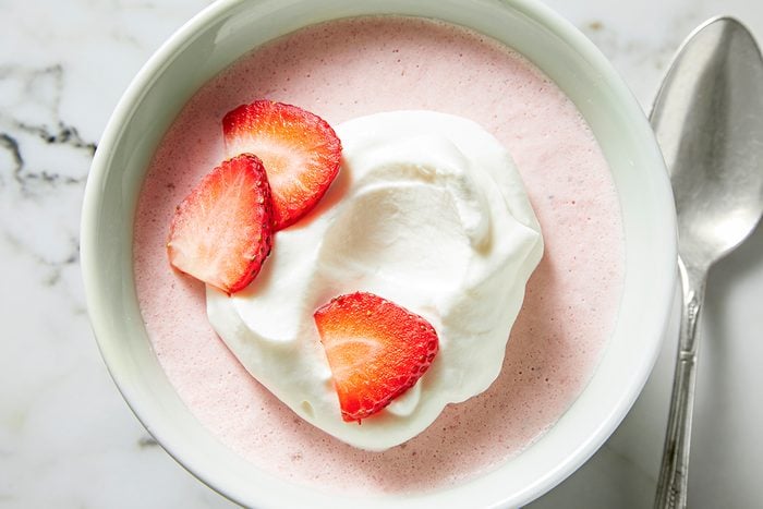 A bowl of pink strawberry mousse topped with a dollop of whipped cream and three fresh strawberry slices. A silver spoon lies beside the bowl on a marble surface.