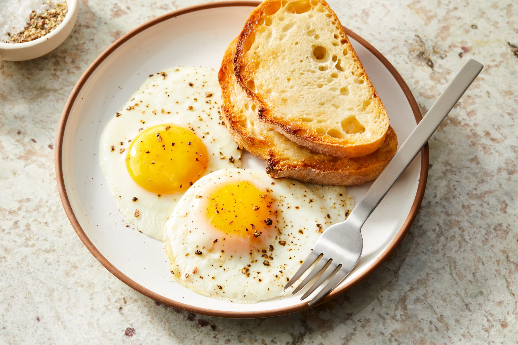 Close shot of Sunny Side Up Eggs; Served hot on a palte with breads slices; and fork; marble surface;