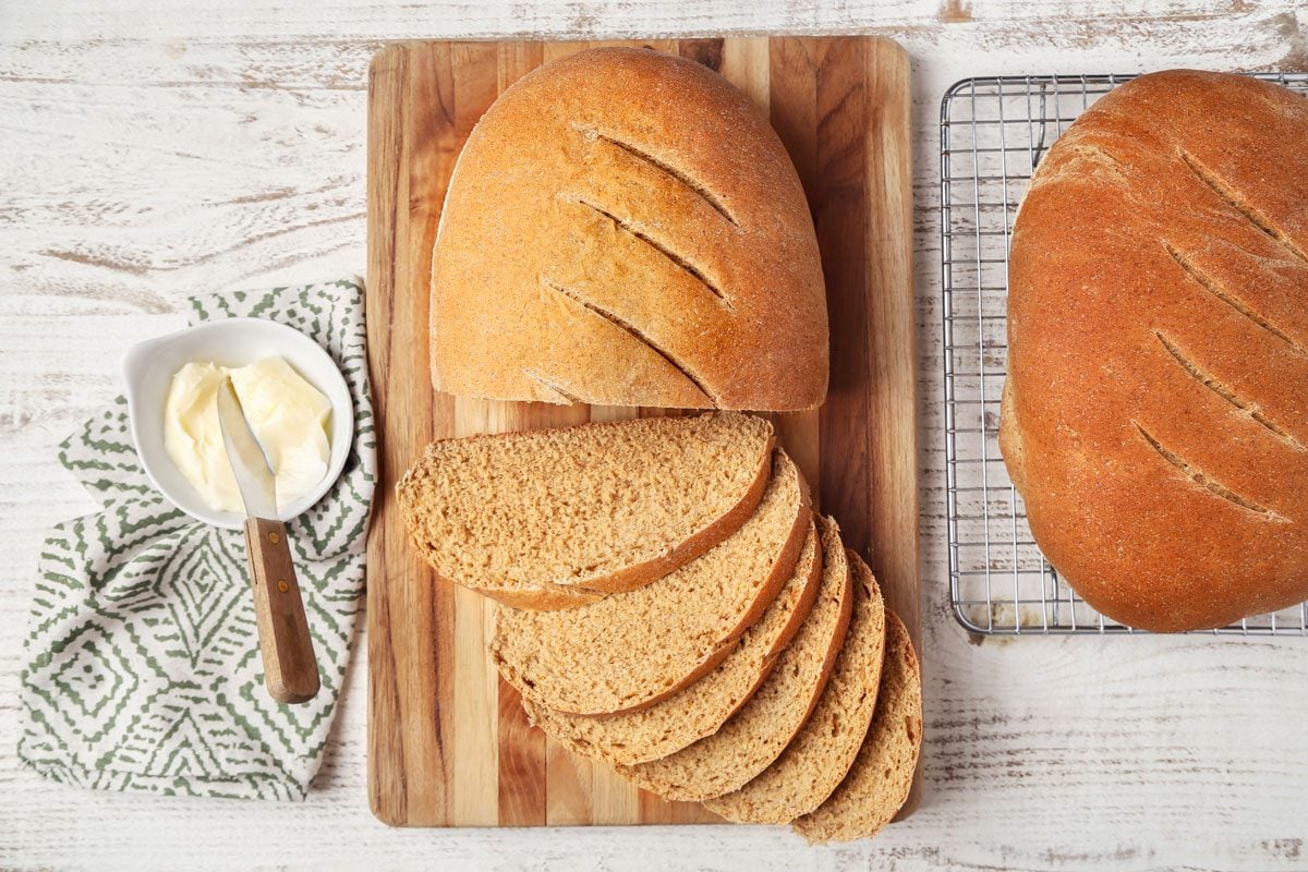 overhead shot of Swedish Limpa Bread, there are two loaves of Swedish Limpa Bread, one whole loaf is on a wire rack and the other is cut and displayed on a wooden cutting board, to the left of the cutting board, there is a white bowl with butter and a butter knife, the background is a white wood surface