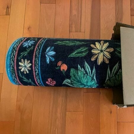 Black rug with floral and leaf design rolled up emerging from a box on a wood floor