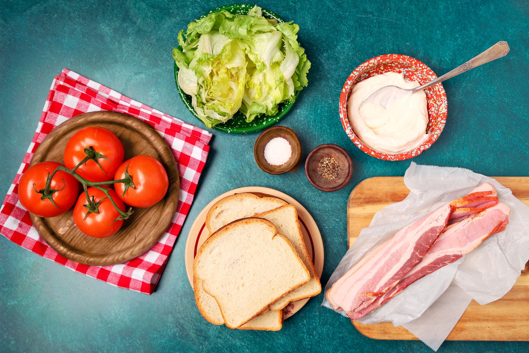 overhead shot of various ingredients for a classic BLT sandwich laid out on a teal background; there are four vine-ripened tomatoes on a wooden platter with a red and white checkered napkin; next to it there is a plate of white bread; on the right, three strips of bacon are on parchment paper resting on a wooden cutting board